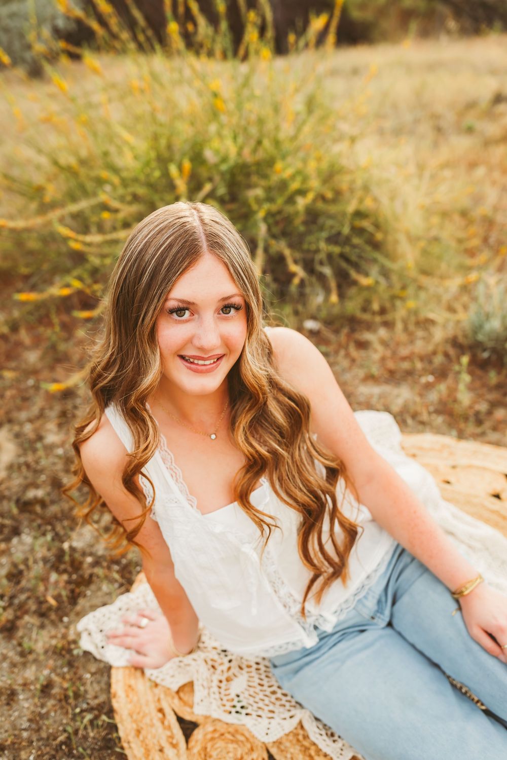 Young woman with wavy hair smiling in white lace top and jeans, seated outdoors in golden field.