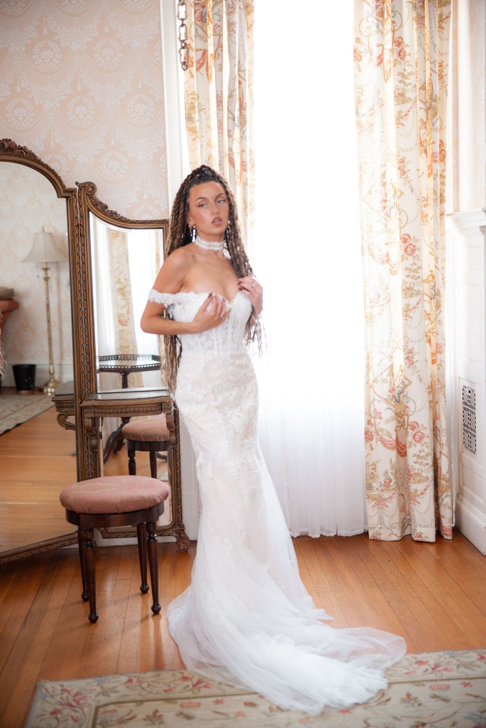 Bride in lace mermaid wedding gown with dreadlocks stands by ornate mirror in elegant vintage room.
