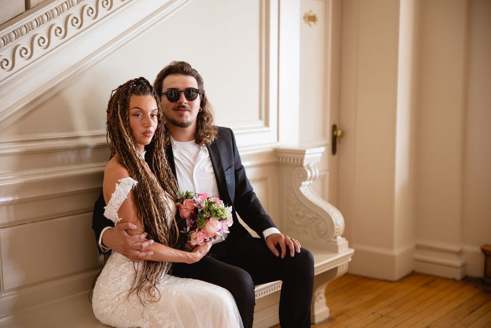 Stylish couple in wedding attire sitting together on bench indoors, groom wearing sunglasses.