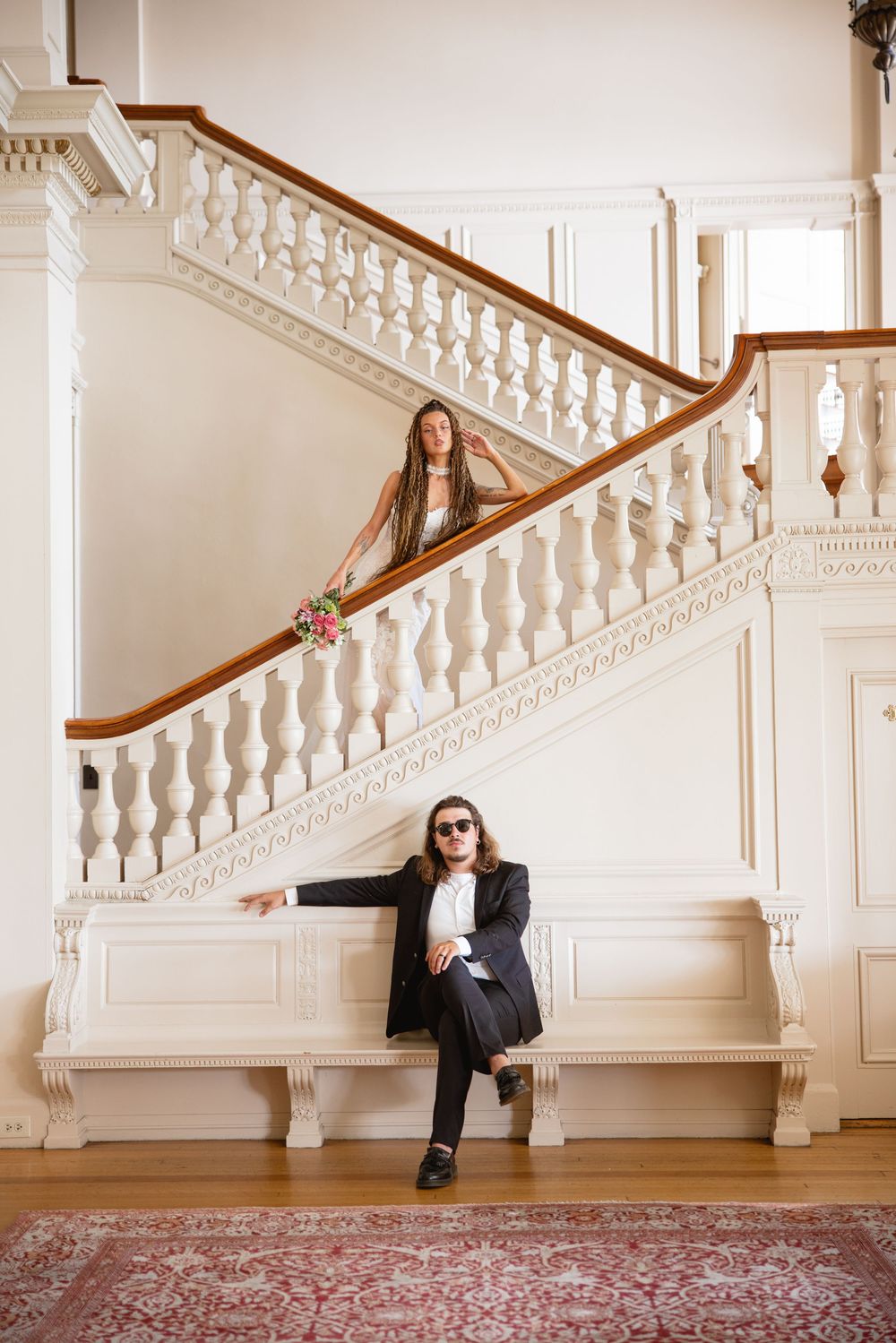 Bride descends grand white staircase with bouquet while groom in black suit sits casually below.