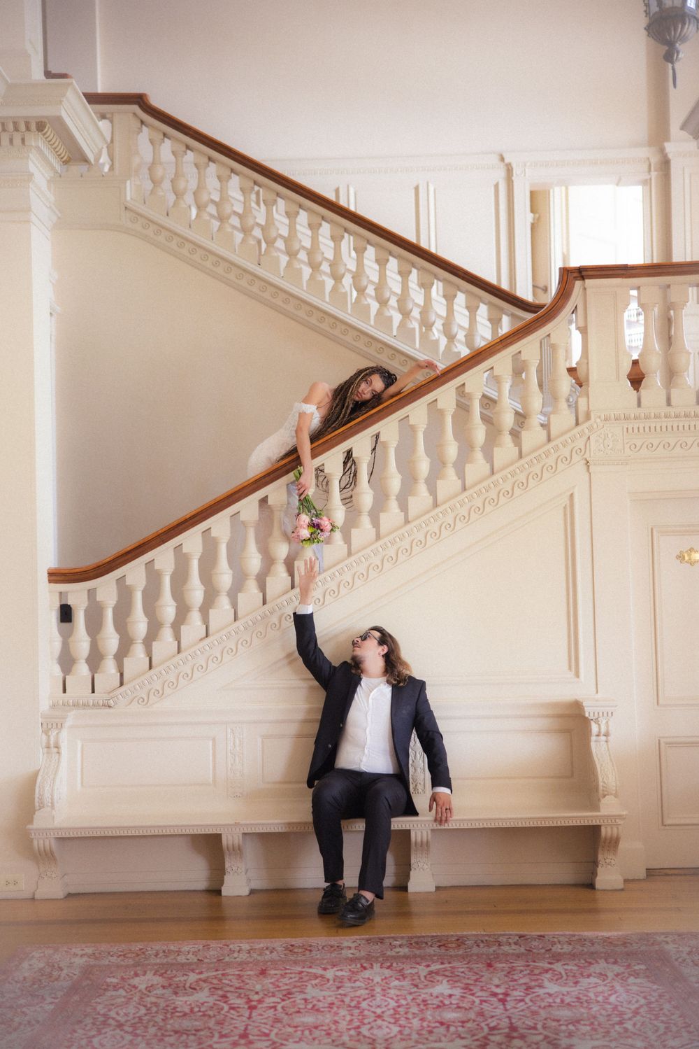 Groom reaches up toward bride on white staircase as she lowers her floral bouquet to him.