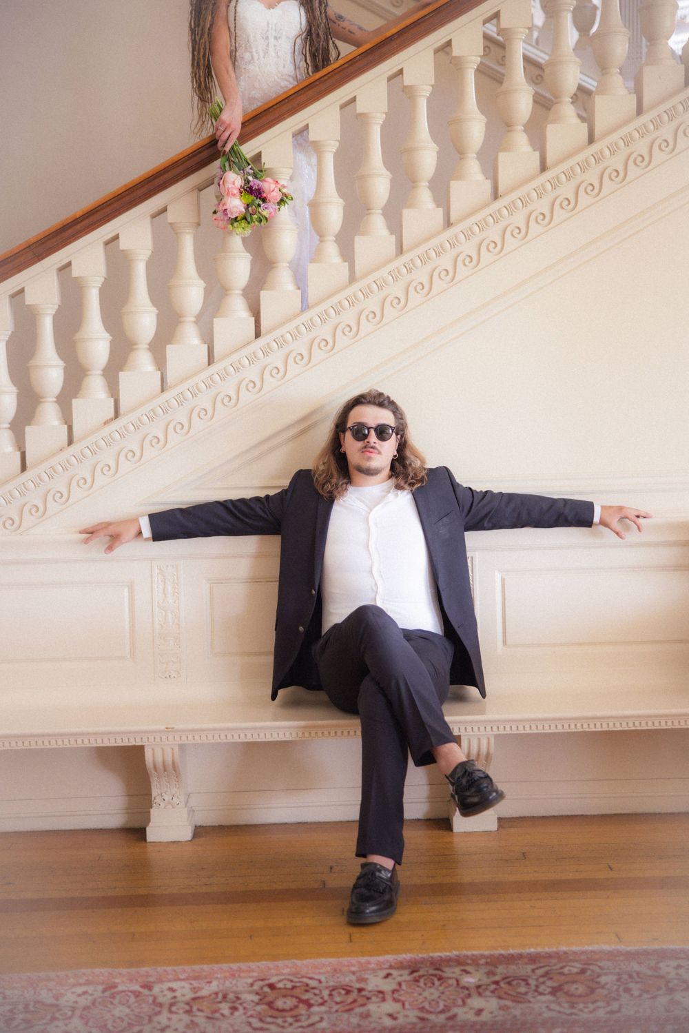 Groom in black suit and sunglasses sits on bench with arms spread wide beneath grand staircase.