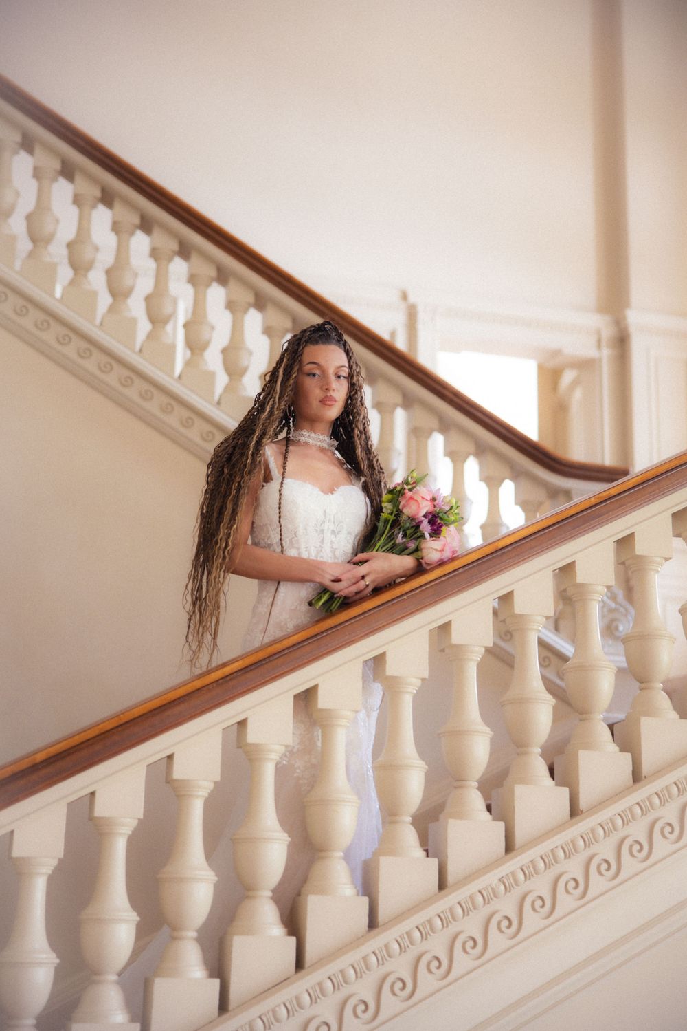 Bride with long dreadlocks in white lace gown holds pink bouquet on elegant white staircase.