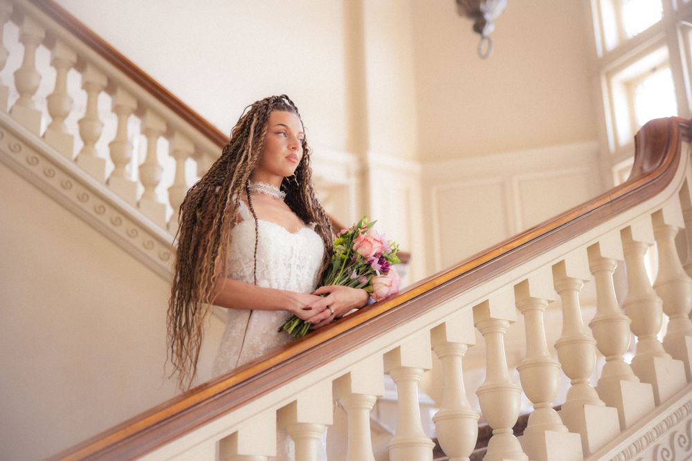 Bride with long dreadlocks holds pink floral bouquet while standing on ornate white staircase railing.