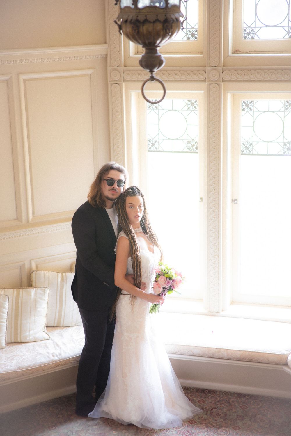 Bride and groom pose together by stained glass windows, she holds bouquet in lace mermaid gown.