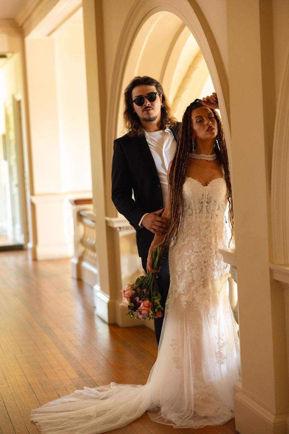 Stylish couple in wedding attire pose beneath elegant arched columns in ornate historic venue hallway.