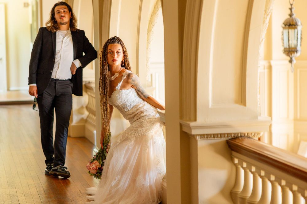 Bride in lace gown and groom in black suit walk elegant corridor during wedding photoshoot.