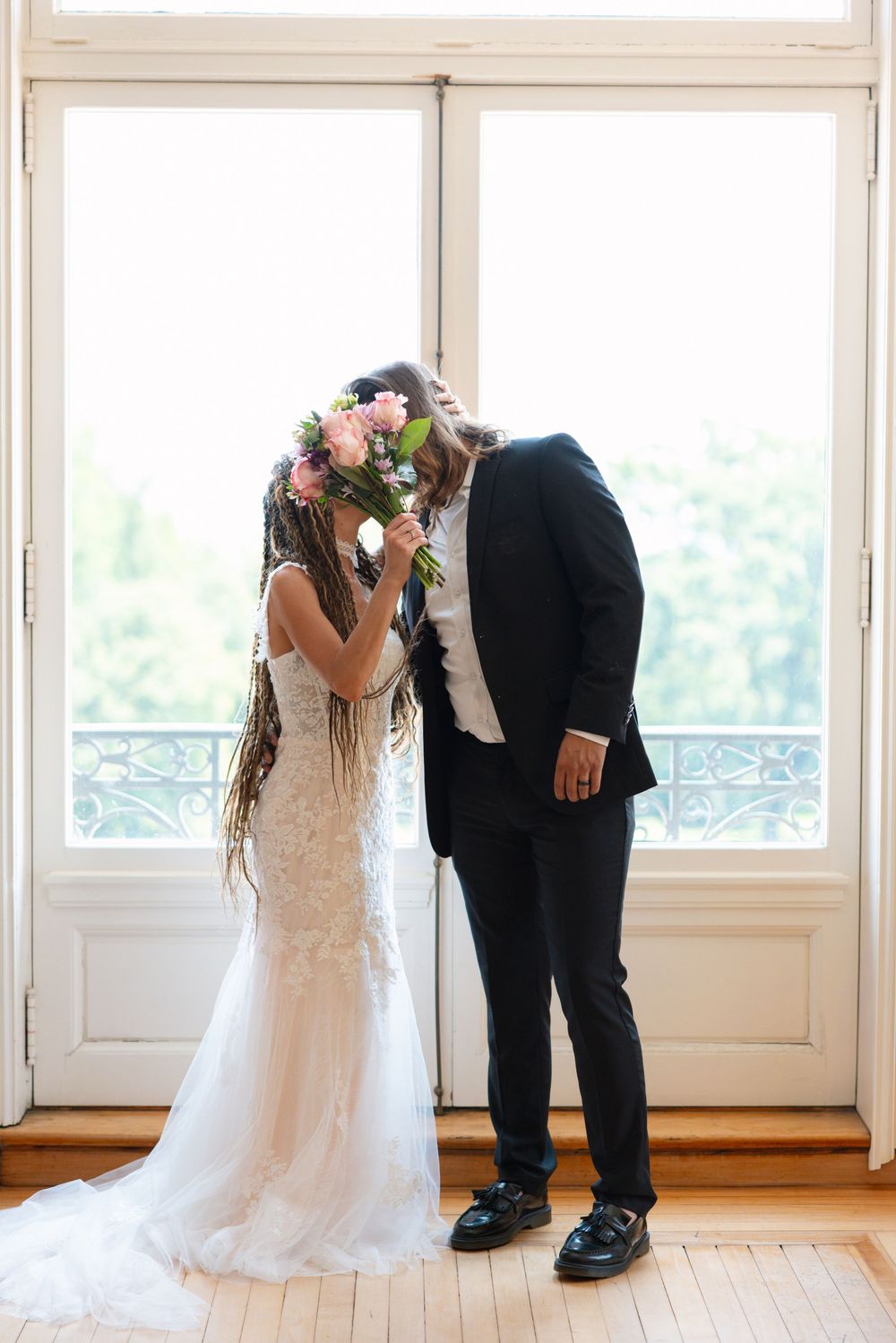 Bride and groom kiss behind pink floral bouquet standing by bright white French doors.