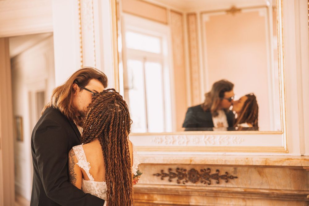 Newlyweds sharing intimate kiss reflected in ornate gold mirror in warmly lit elegant room.