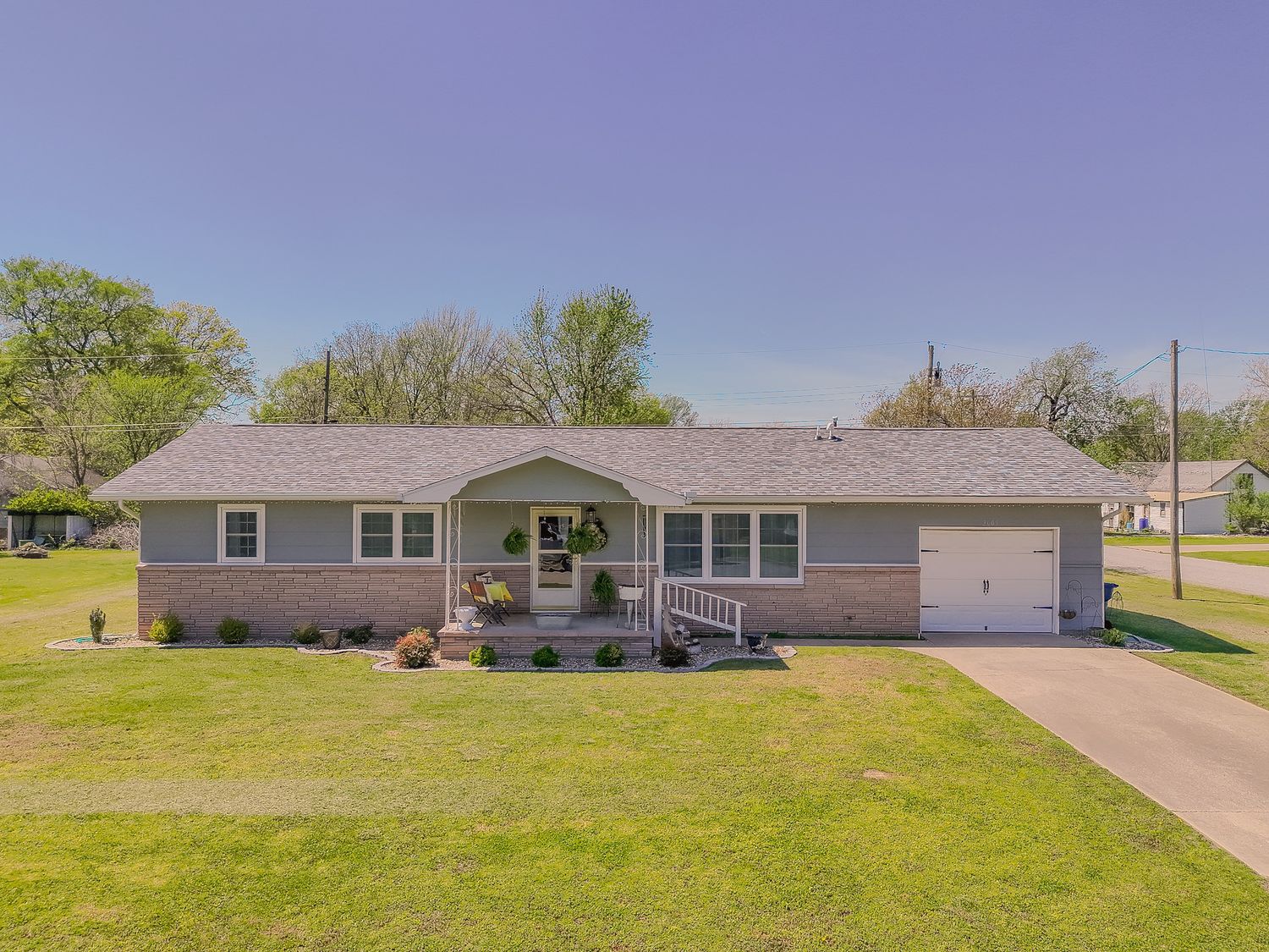 Ranch-style home with gray siding, attached garage, covered porch, and lush green lawn on a sunny day.