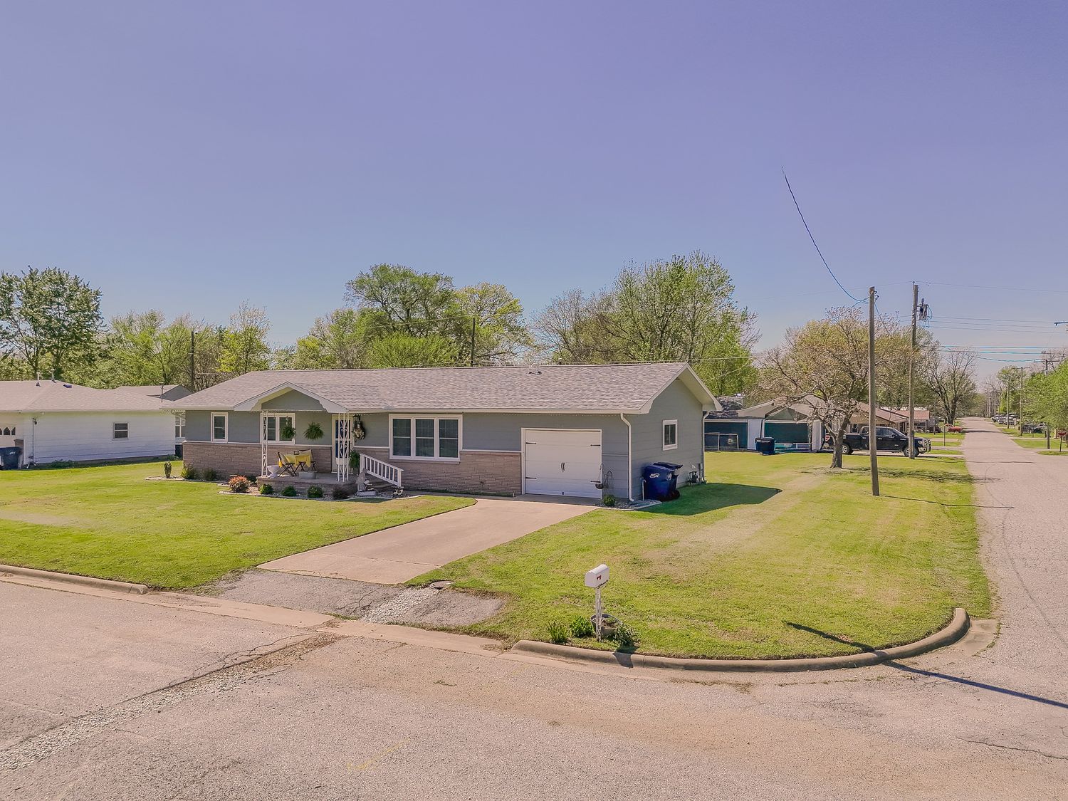 Brick ranch-style home with attached garage, green lawn, and tree-lined street on a sunny day.