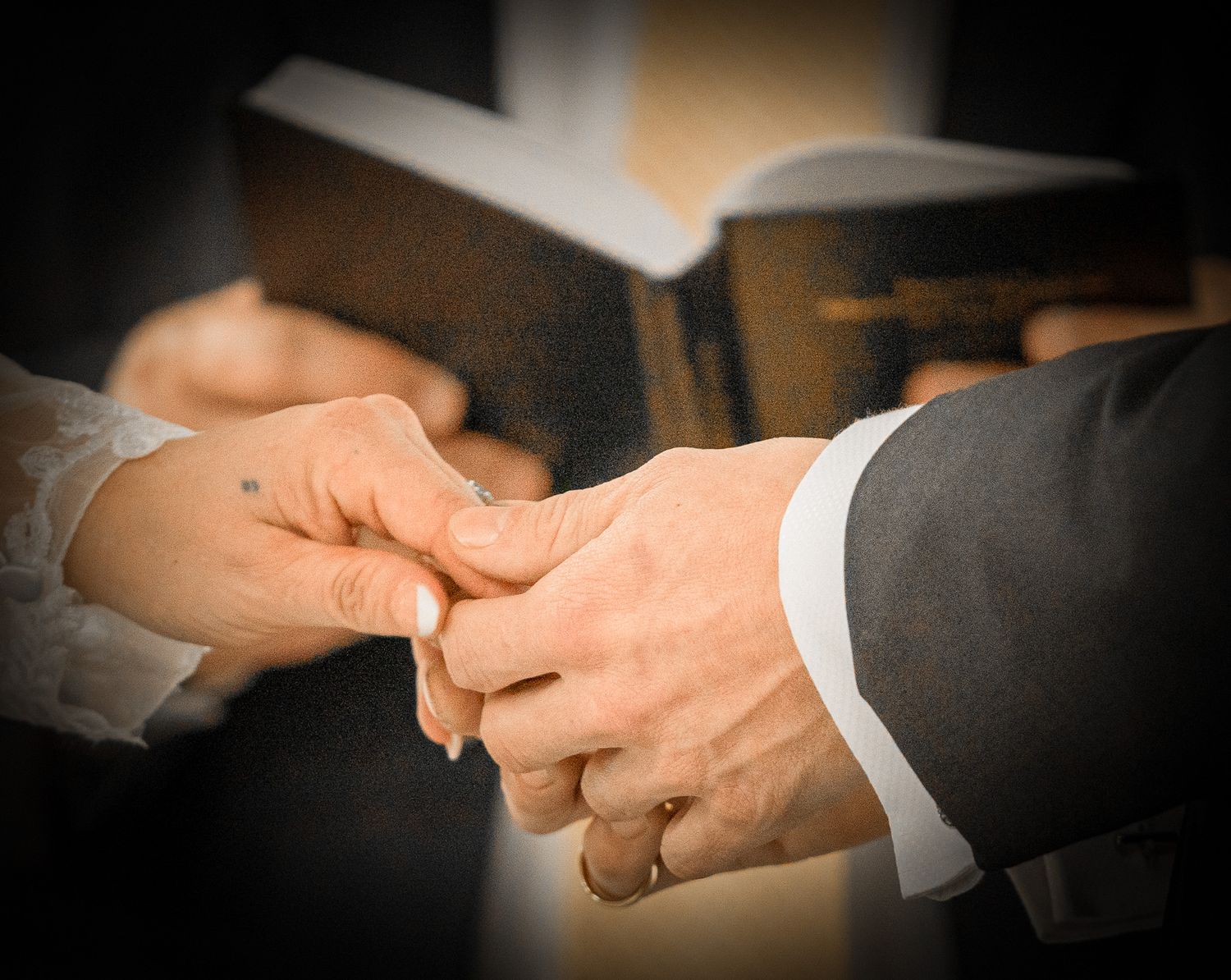Couple exchanging wedding rings during a ceremony, with a Bible held by an officiant in the background.