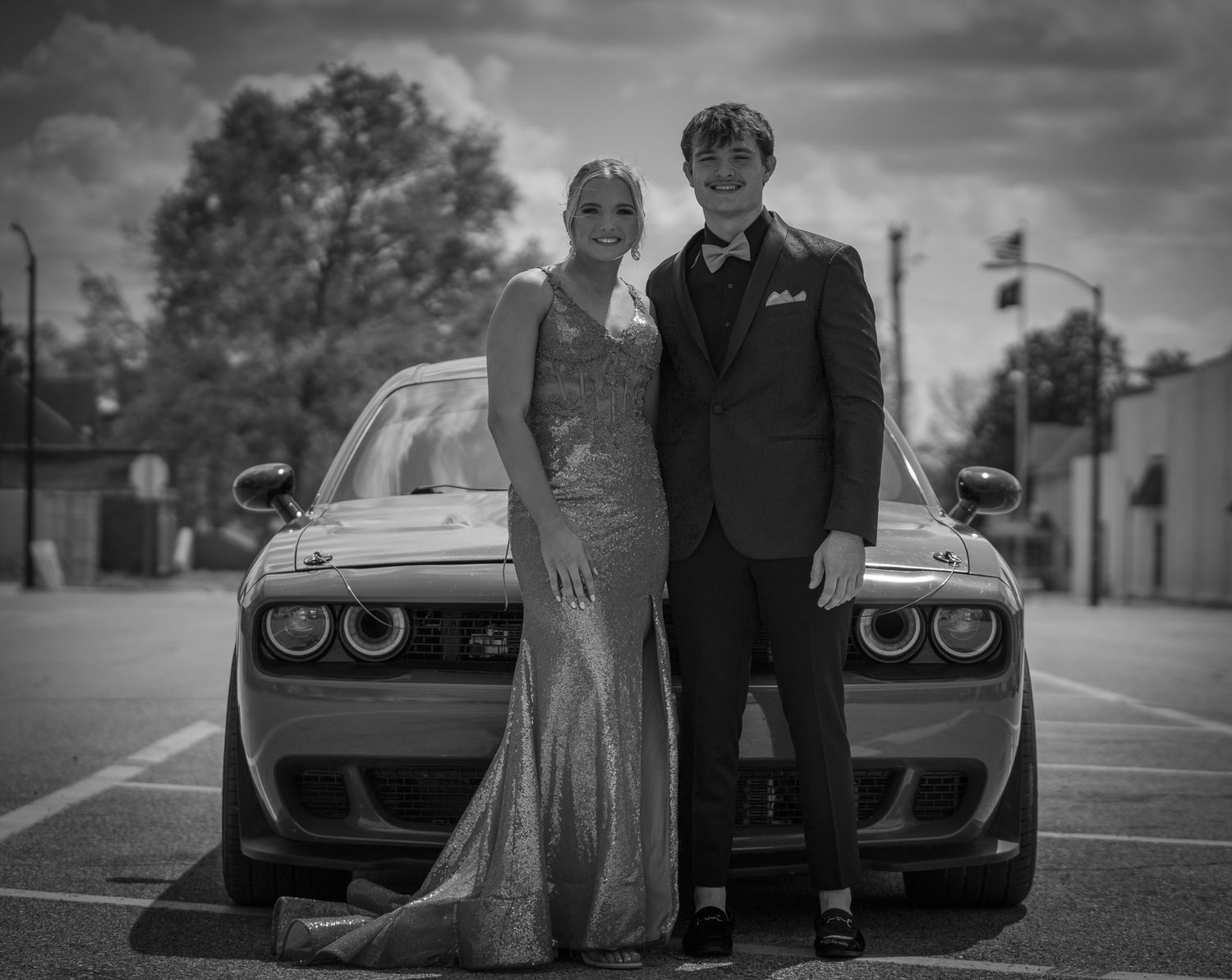 A couple in formal prom attire poses in front of a Dodge Challenger in a black and white photo.