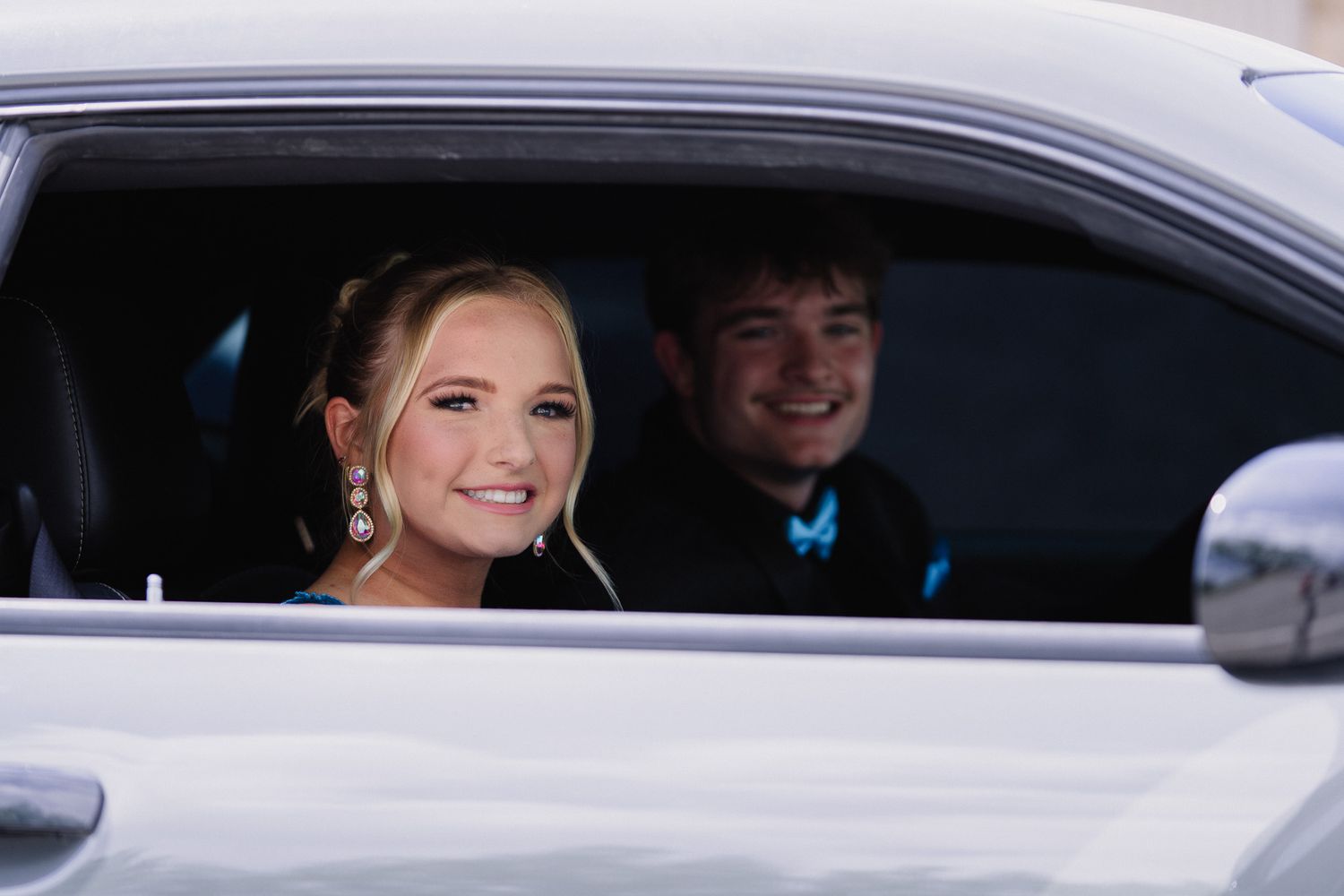 A smiling young couple dressed in formal attire sitting inside a white car, likely heading to prom.