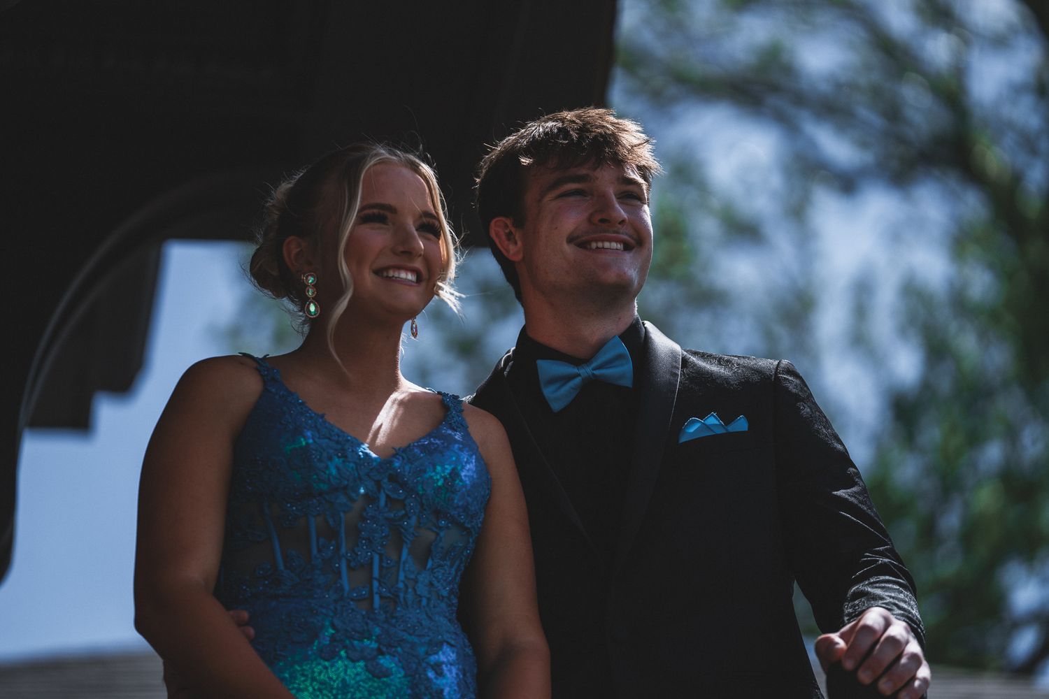 A smiling couple dressed in prom attire, woman in blue gown and man in black suit with bow tie, posing outdoors.