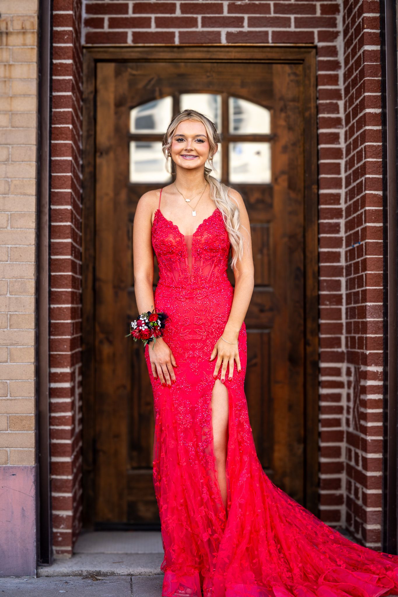 Young woman in a red lace mermaid prom gown with a floral wrist corsage, posing in front of a wooden door.
