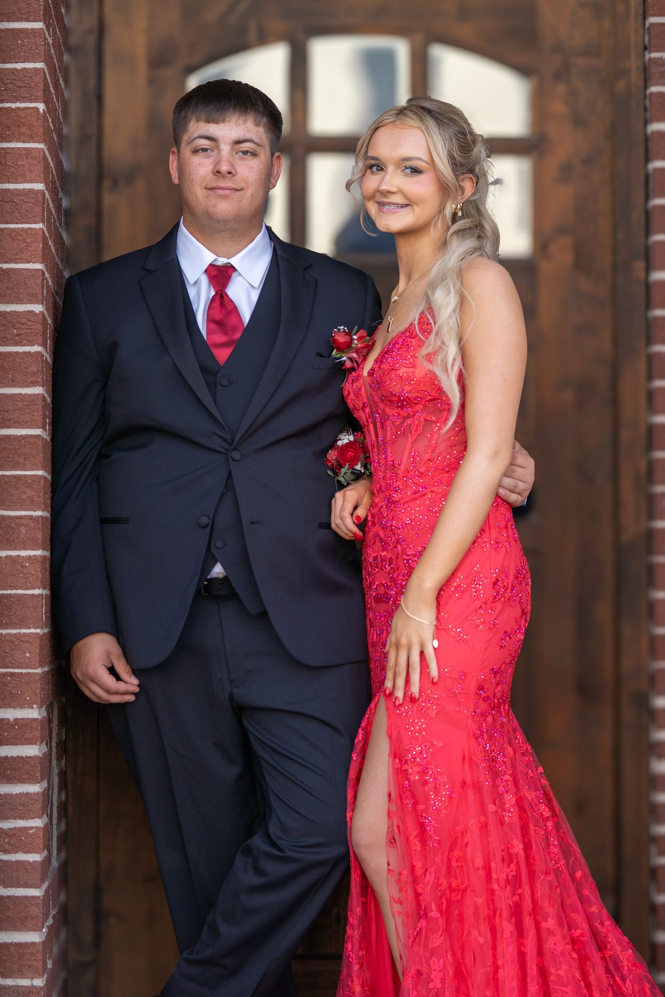 A young couple dressed in formal prom attire, him in a navy suit and her in a red lace gown, pose by a brick wall.