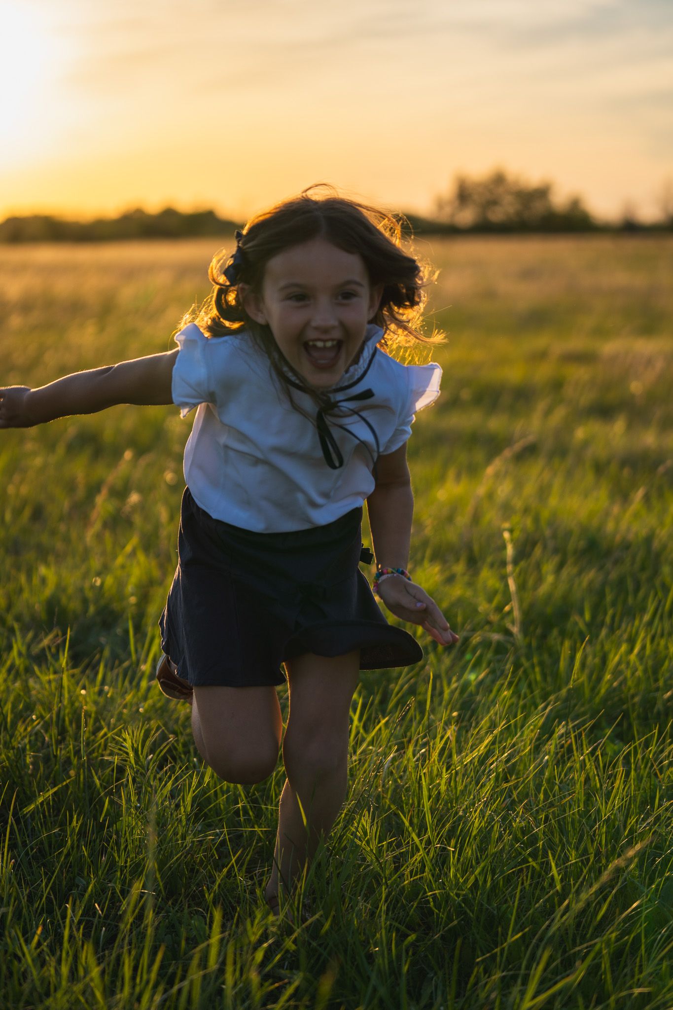 A joyful girl in a white blouse and dark skirt runs through a green field at golden sunset.