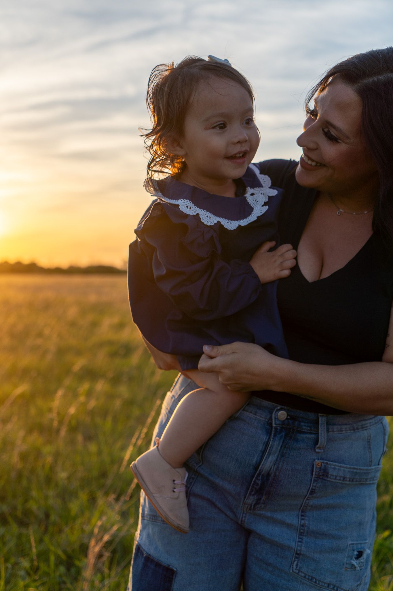A mother holds her smiling toddler in a field at golden sunset, both wearing dark outfits in a heartwarming outdoor portrait.