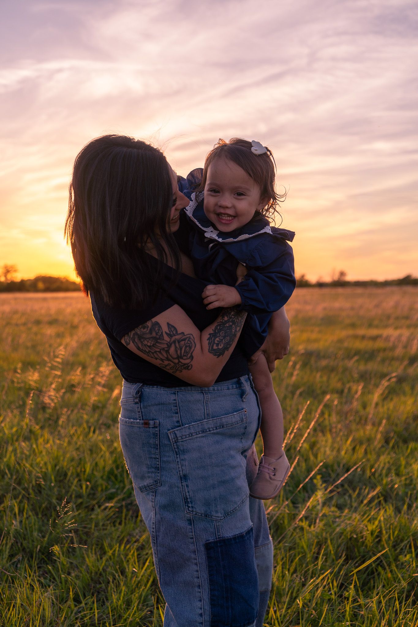A tattooed mother holds her smiling toddler in a golden sunset field, sharing a tender moment outdoors.
