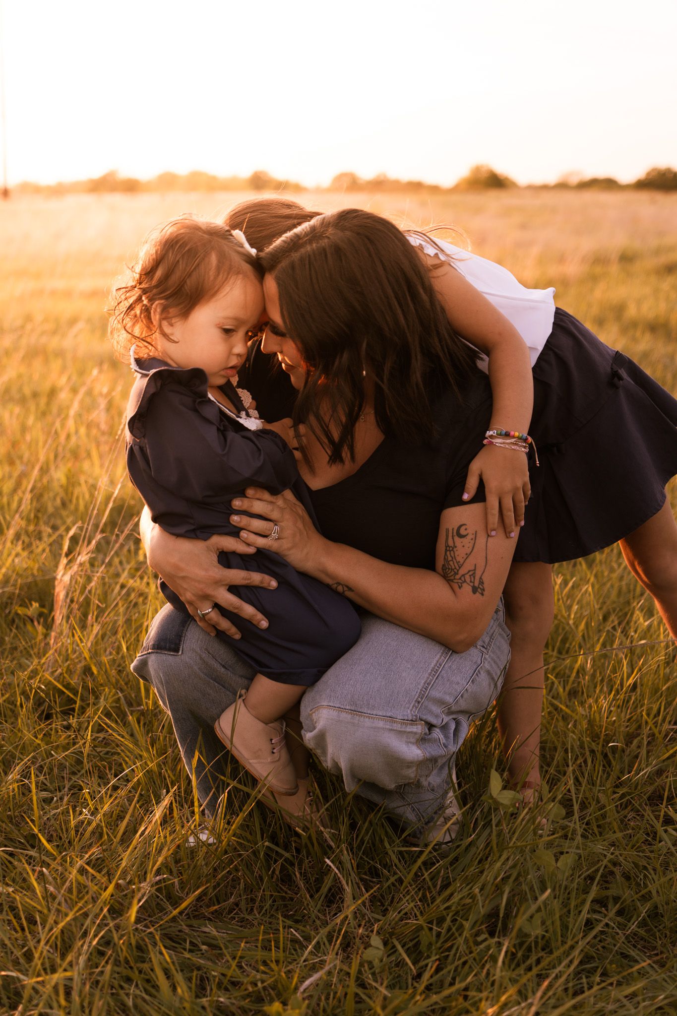 A mother tenderly embraces her toddler in a golden sunlit field at sunset, touching foreheads in a loving moment.