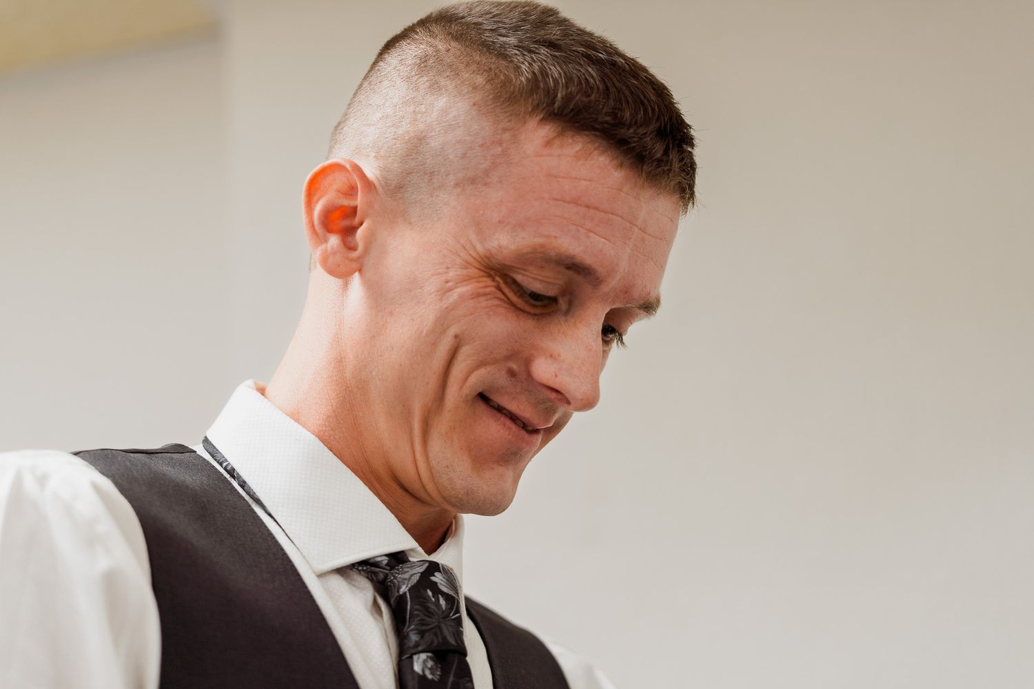 A man in a white dress shirt, dark vest, and patterned tie looks down with a slight smile.