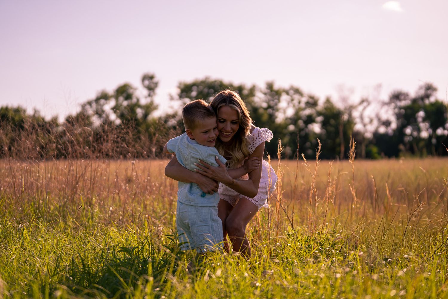 A mother and child share a warm embrace in a sunlit green meadow with trees in the background.