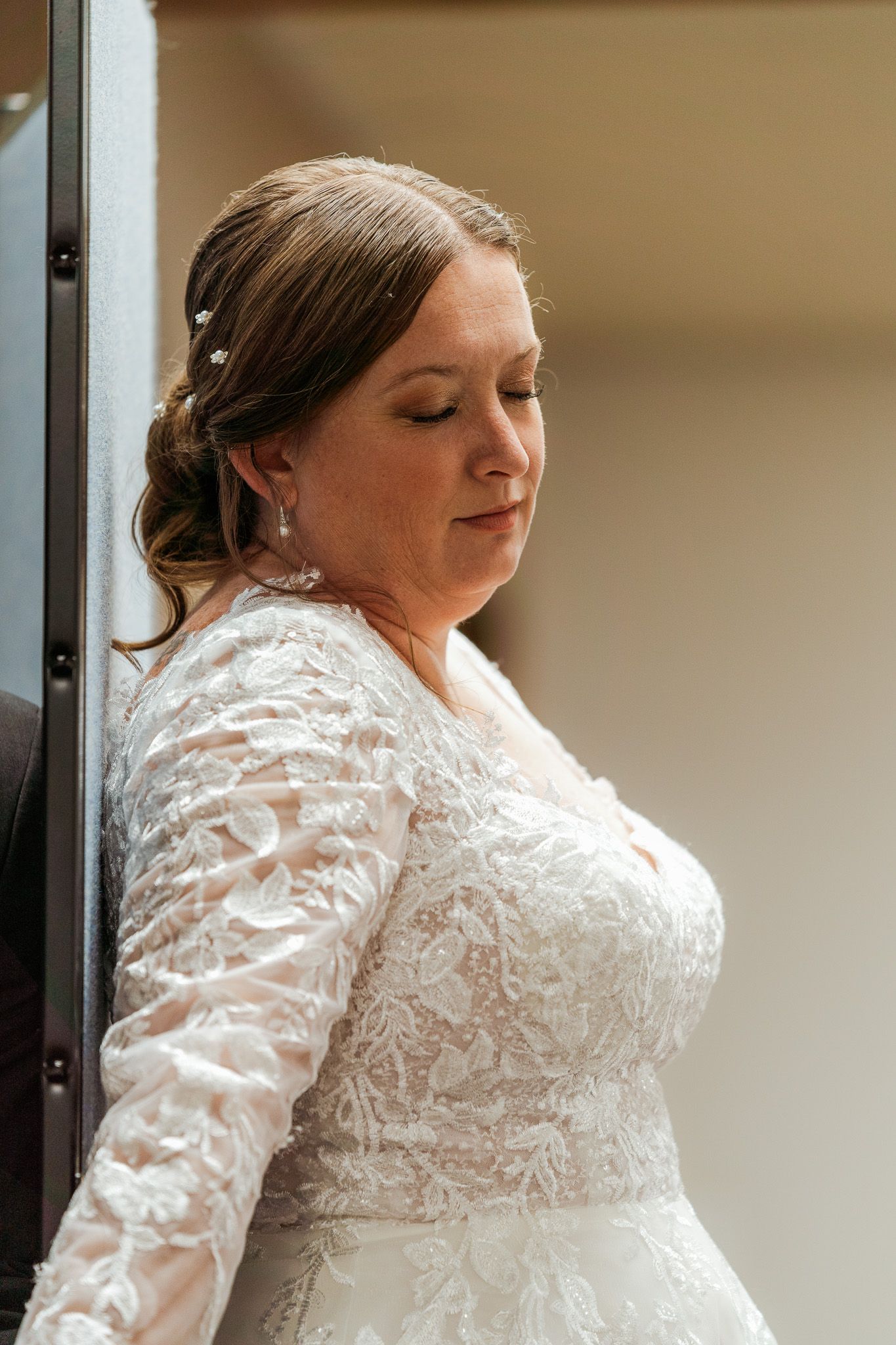 Bride in long-sleeve lace wedding gown with hair accessories, eyes closed, leaning against wall in elegant bridal portrait.