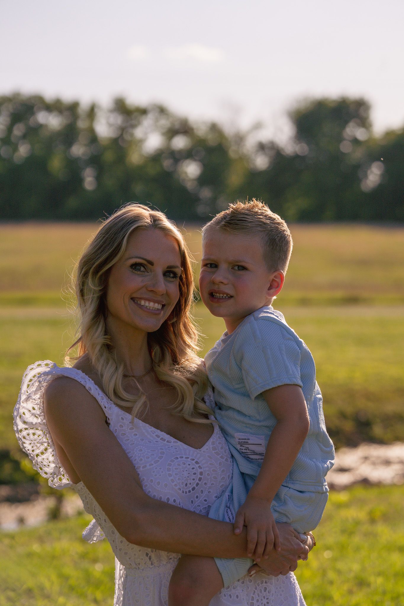 A smiling blonde woman in a white dress holds a young boy in a light blue shirt outdoors in a sunny field.