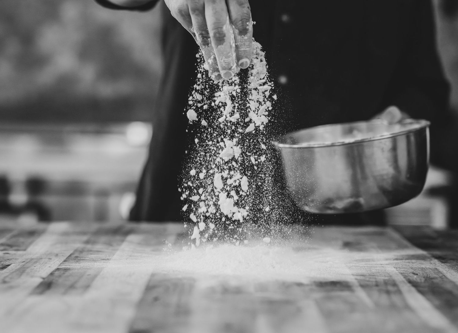 Black and white photo of a baker dusting flour onto a wooden surface beside a metal mixing bowl.