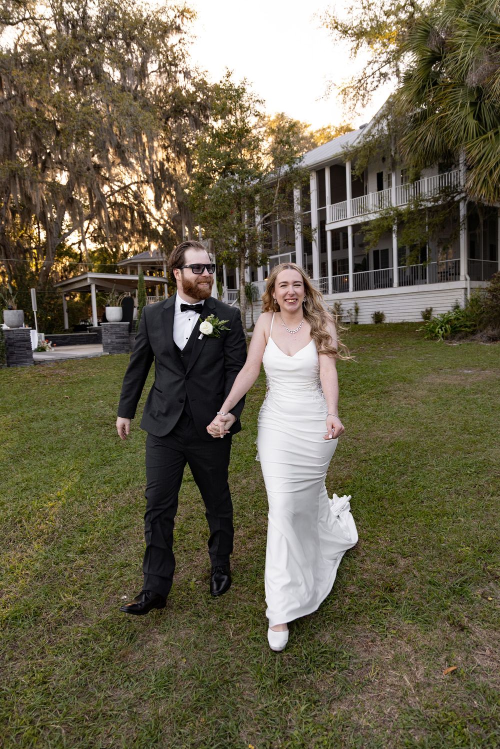 Bride and groom walk hand-in-hand on green lawn outside elegant Southern venue draped in Spanish moss.