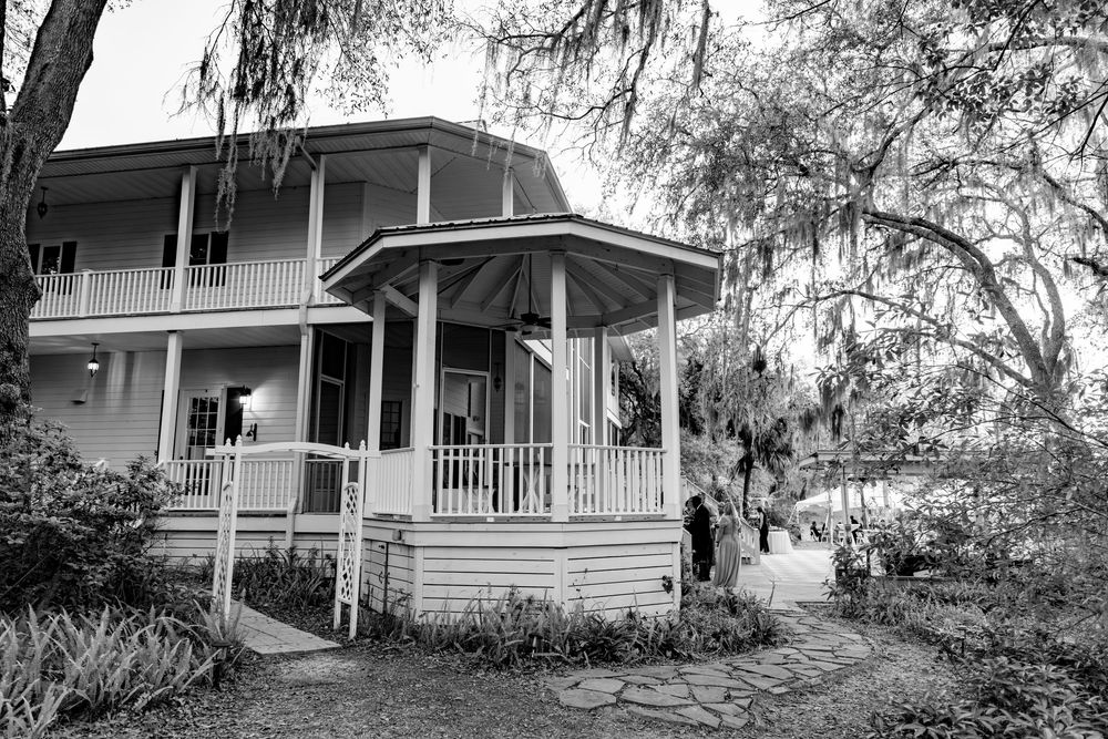 Black and white photo of a charming two-story Southern home with wrap-around porch and oak trees.
