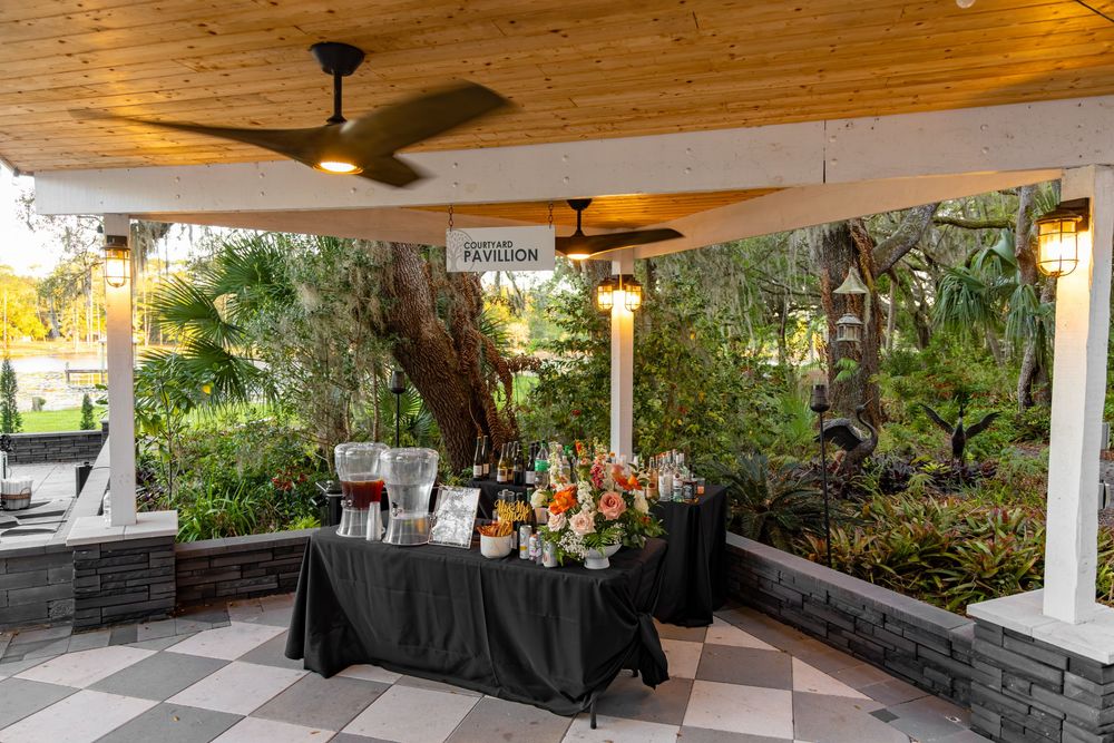 Elegant outdoor wedding reception setup with black draped buffet table under a covered patio surrounded by lush greenery.