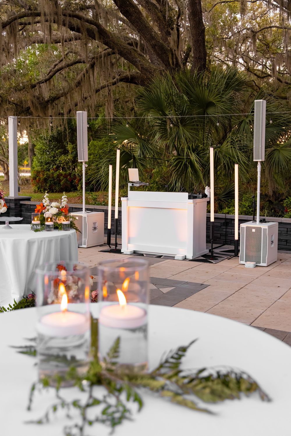 Outdoor wedding DJ setup with white booth, tall speakers, and candlelit tables surrounded by Spanish moss-draped trees.