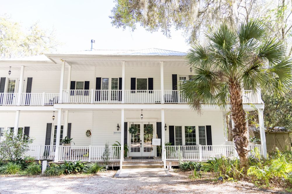 White two-story Southern plantation-style home with black shutters, wrap-around porch, and palm tree in lush setting.
