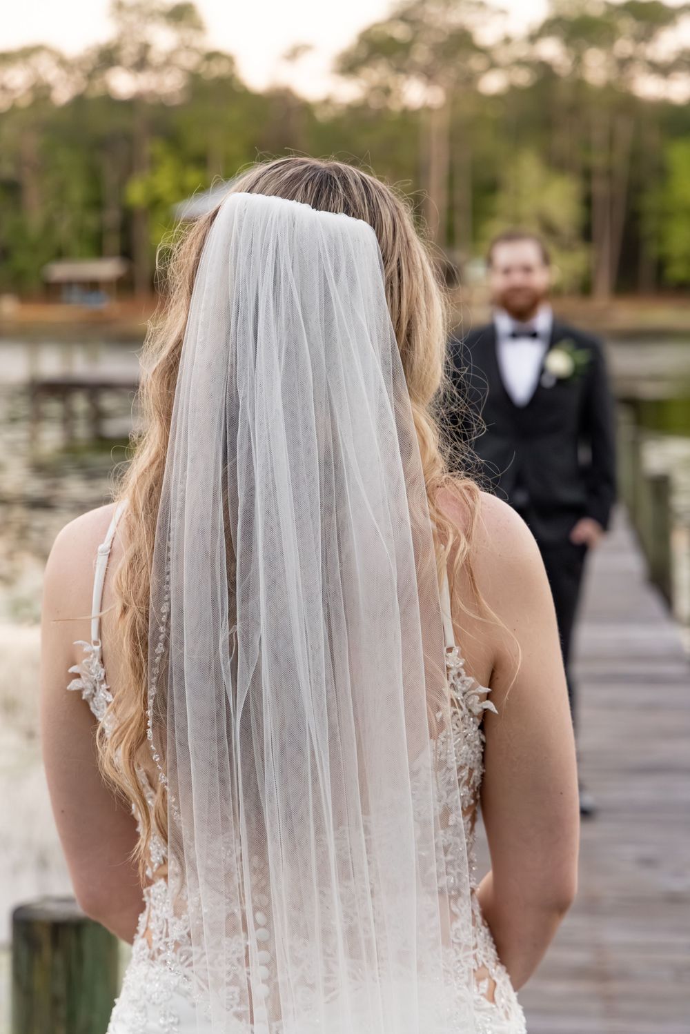 Bride in lace wedding gown and veil stands on dock facing groom in black tuxedo by lakeside.