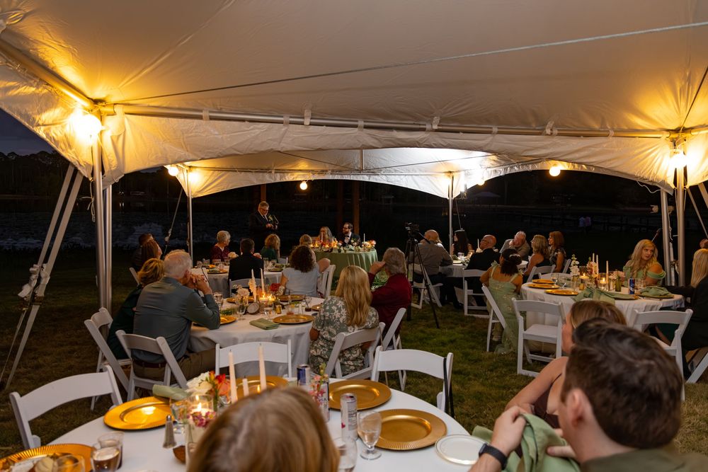 Guests dining under a white tent at an outdoor evening wedding reception with warm string lights.