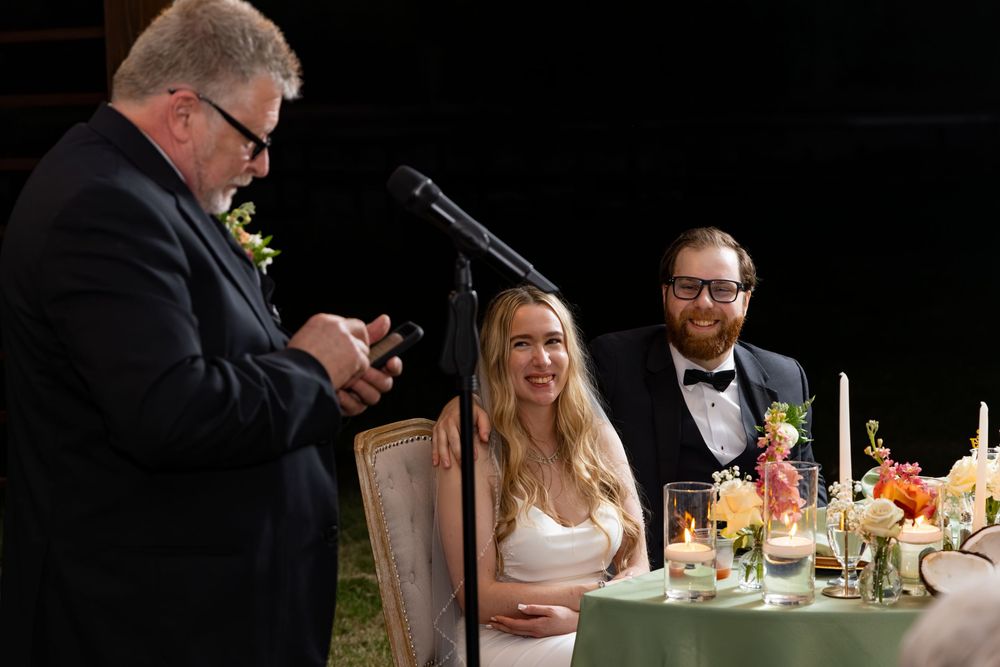Wedding toast scene with man speaking at microphone while bride in white dress and groom in tuxedo smile at reception table.