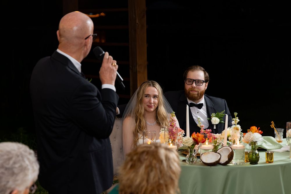 A bald man gives a wedding toast at a reception as the smiling bride in a veil and groom in a tuxedo listen.