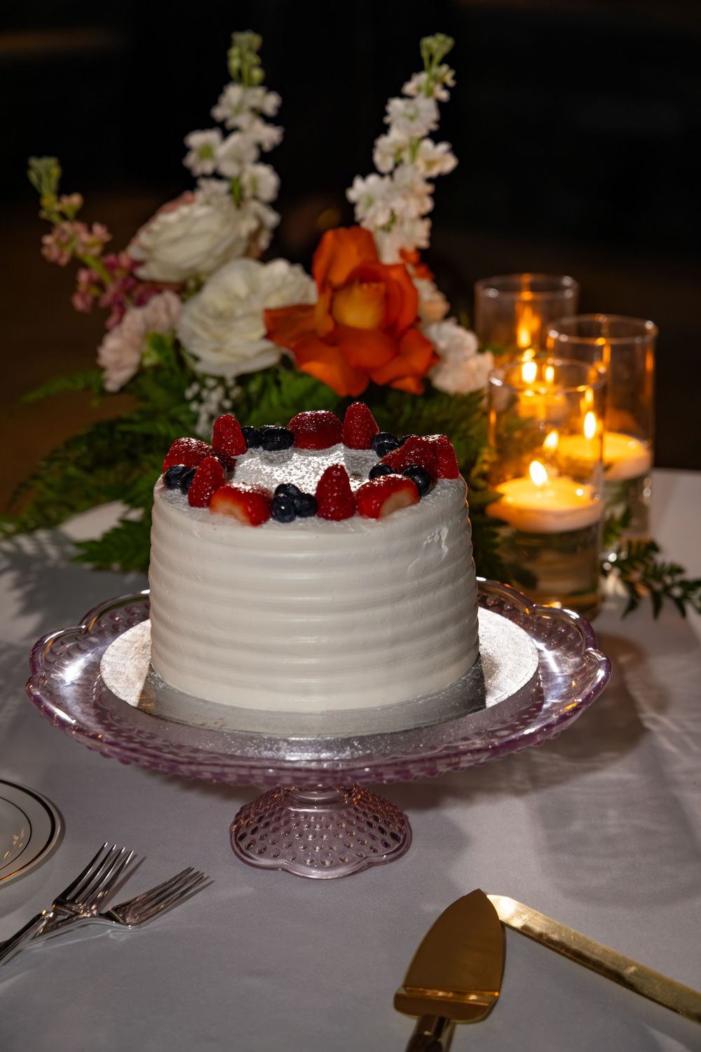 White wedding cake topped with fresh strawberries and blueberries on pink glass stand with floral arrangement and candles.