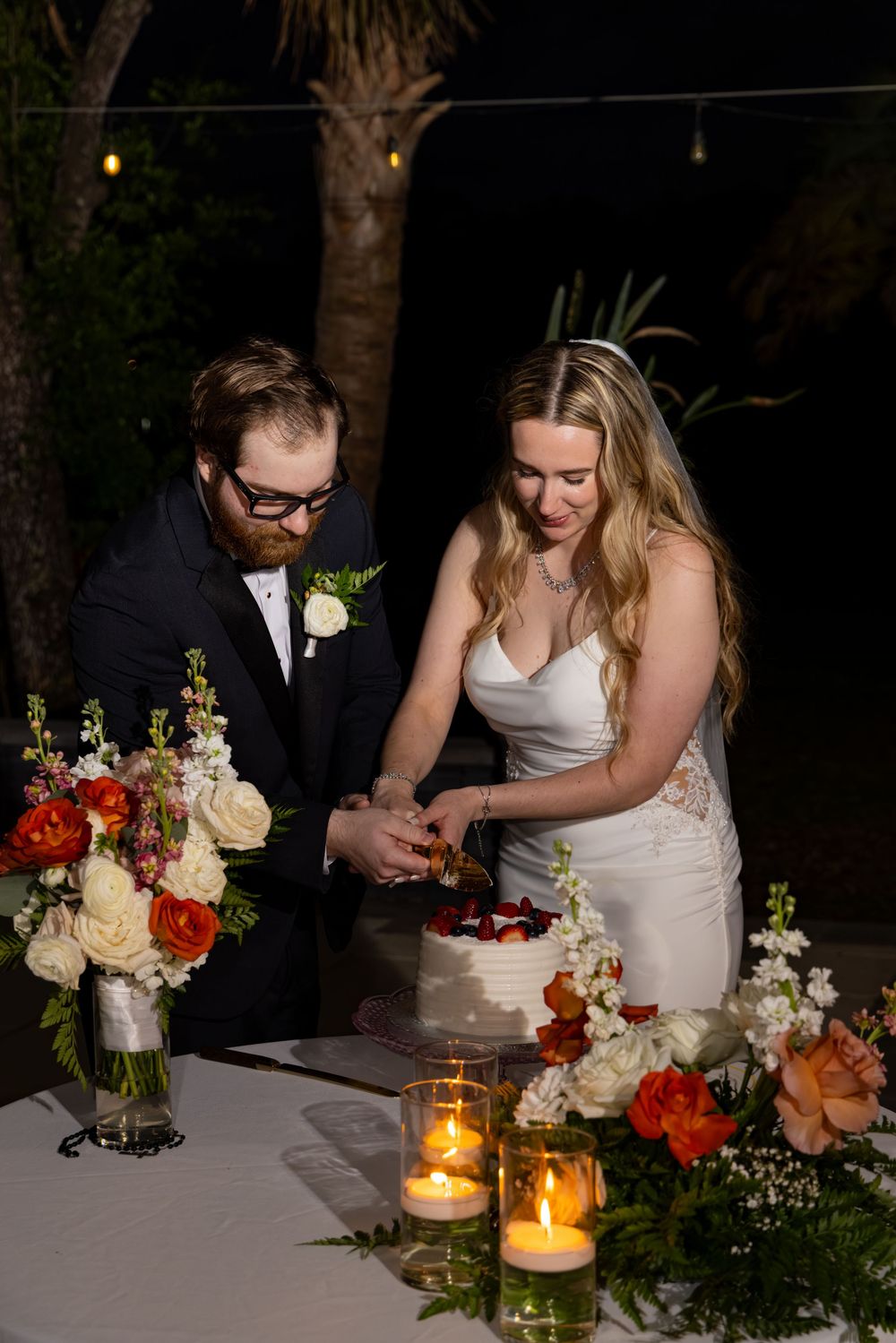 Bride and groom cutting their white wedding cake surrounded by candles and floral arrangements.