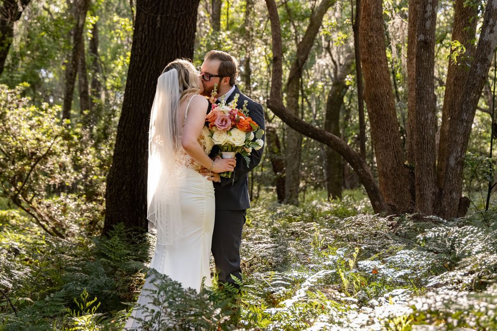 Bride and groom share a tender kiss in a sunlit forest, bride holding a vibrant bouquet.