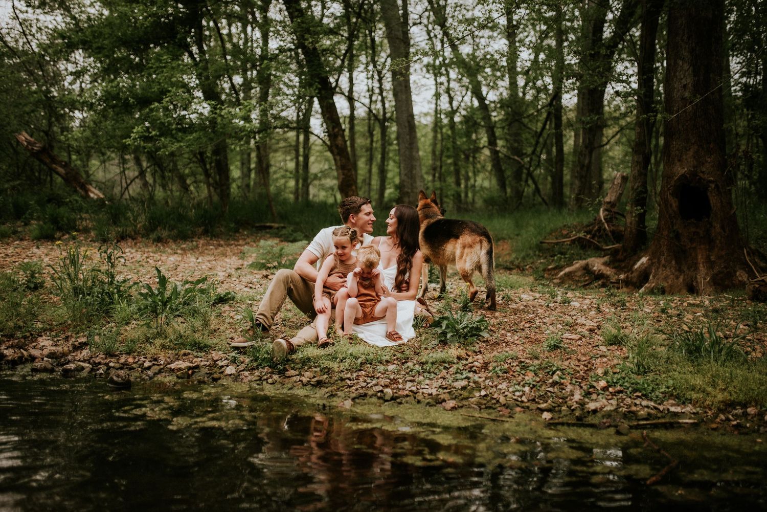 Family sitting creekside as they laugh within each other as photographer stands in the creek to take a photo.