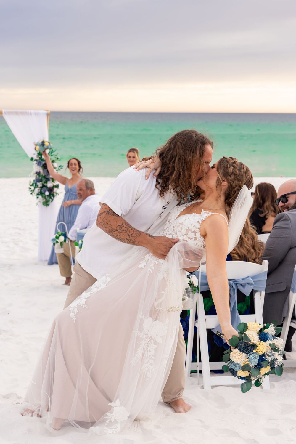 Bride and groom share first kiss at romantic beach wedding ceremony at sunset with wedding party.