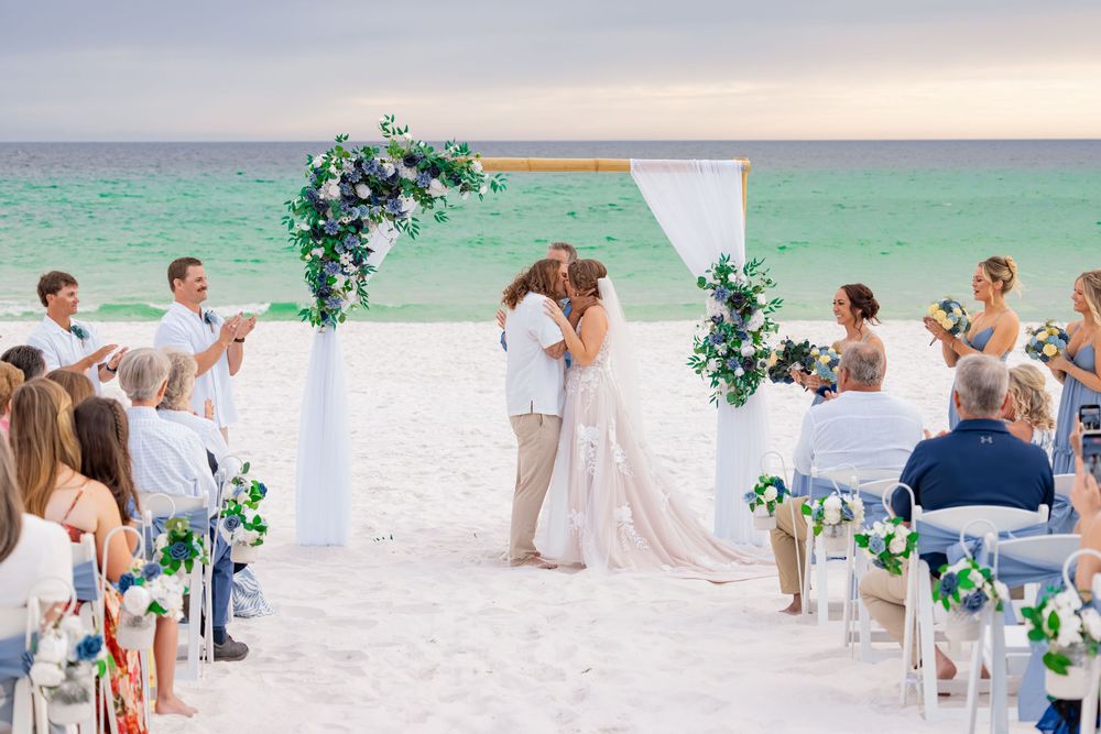 Couple shares first kiss at romantic beach wedding ceremony with blue floral arch and turquoise ocean backdrop.