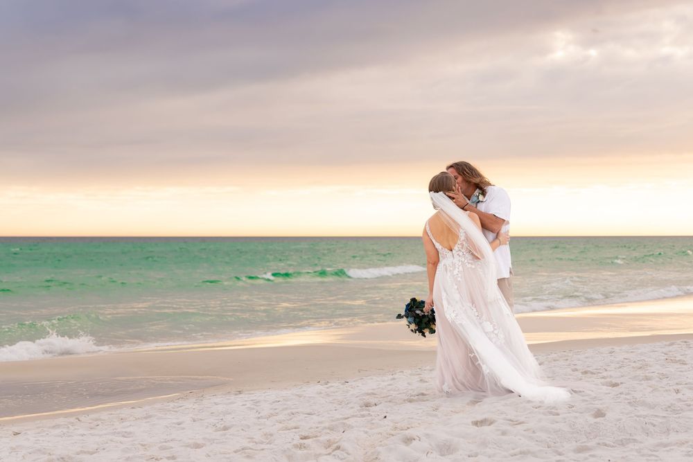 Newlywed couple embracing on white sandy beach at sunset with turquoise ocean in background.