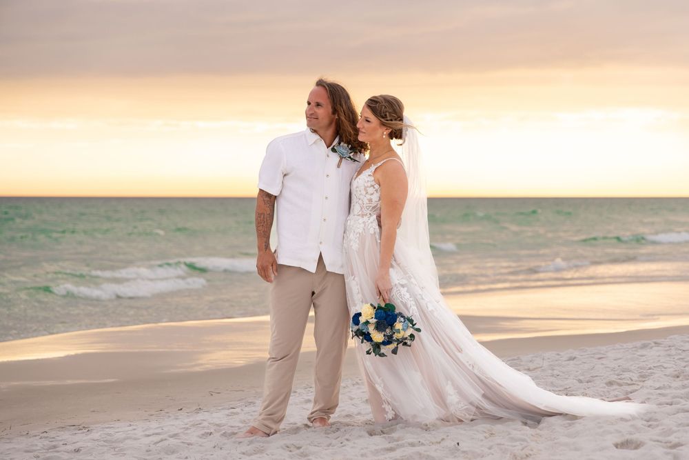 Bride and groom stand barefoot on a sandy beach at sunset, bride holding a blue bouquet in a white gown.