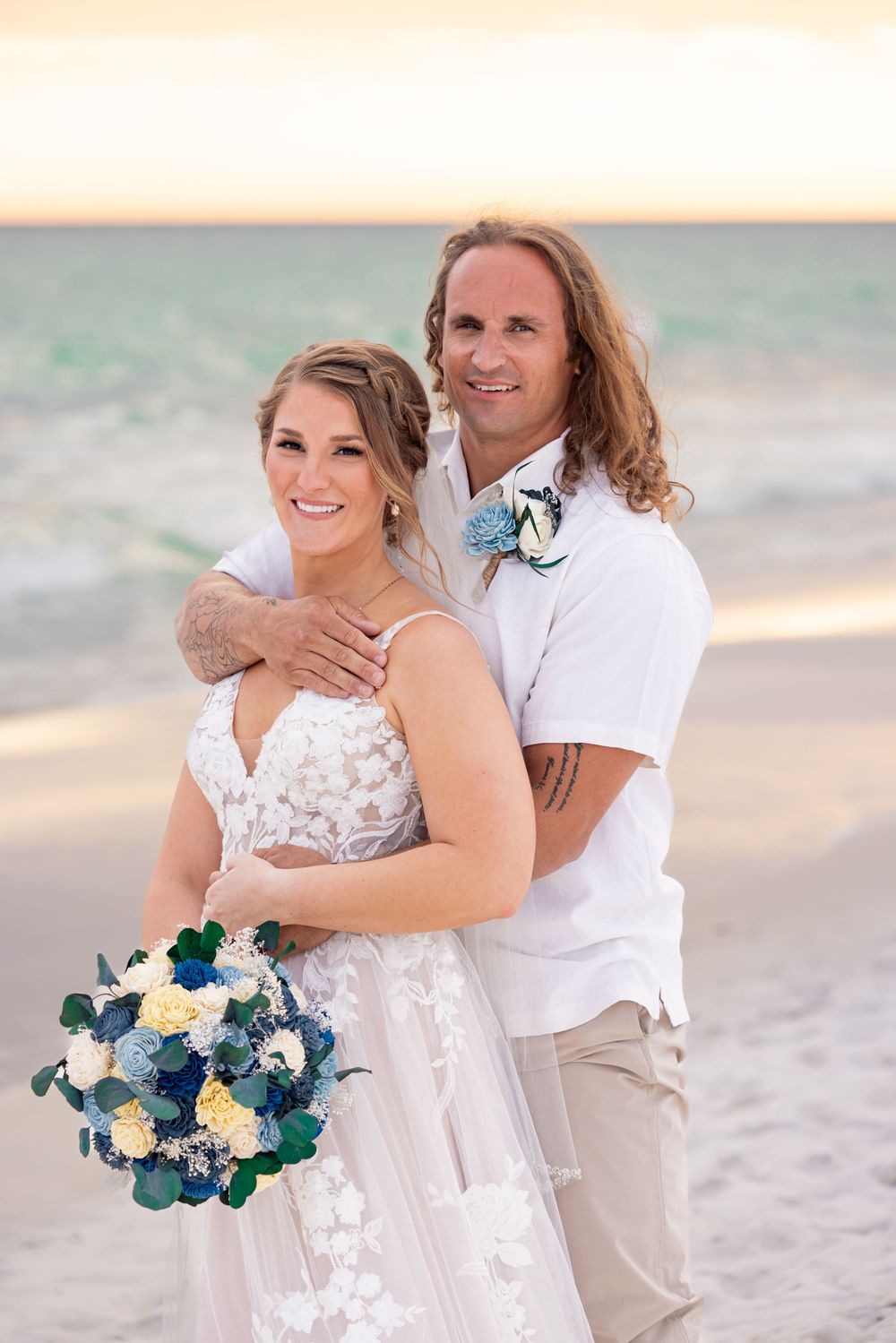 Bride and groom embracing on a white sand beach at sunset, bride holding blue and white bouquet.