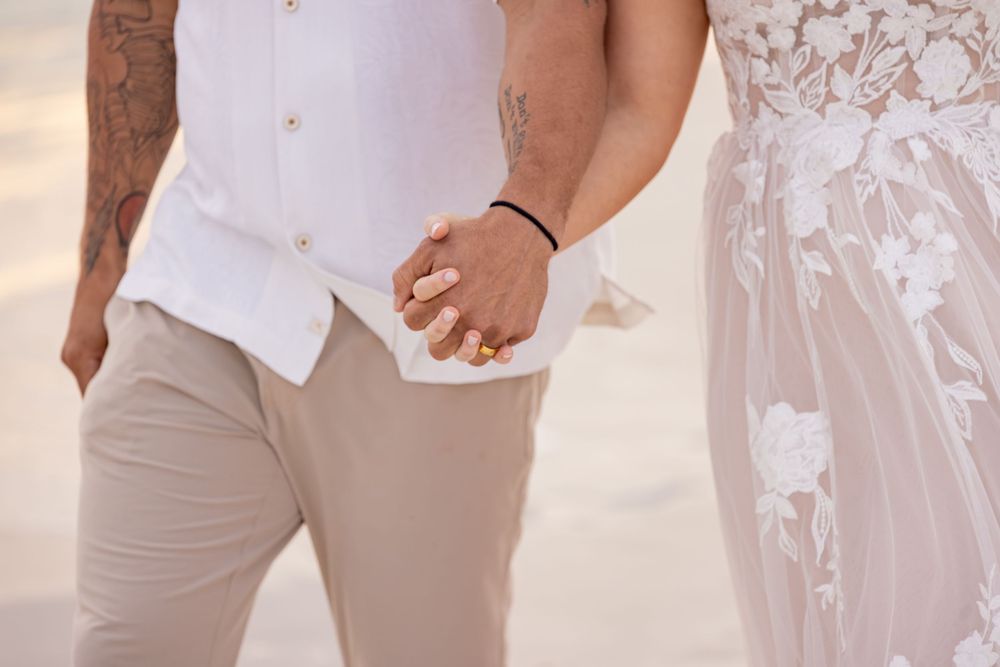 Couple holding hands at beach wedding, groom in white shirt and khakis, bride in lace gown with ring visible.