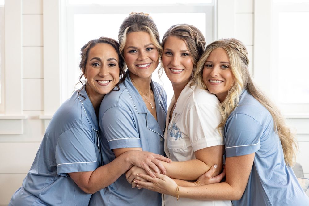 Four bridesmaids in blue robes embrace the bride in white during a bright, elegant wedding preparation photo.